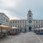 Piazza storica con architettura veneta e gente che passeggia nel sole.