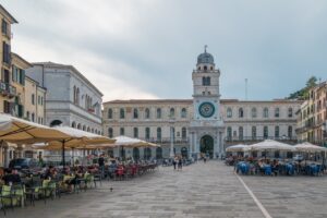 Piazza storica con architettura veneta e gente che passeggia nel sole.